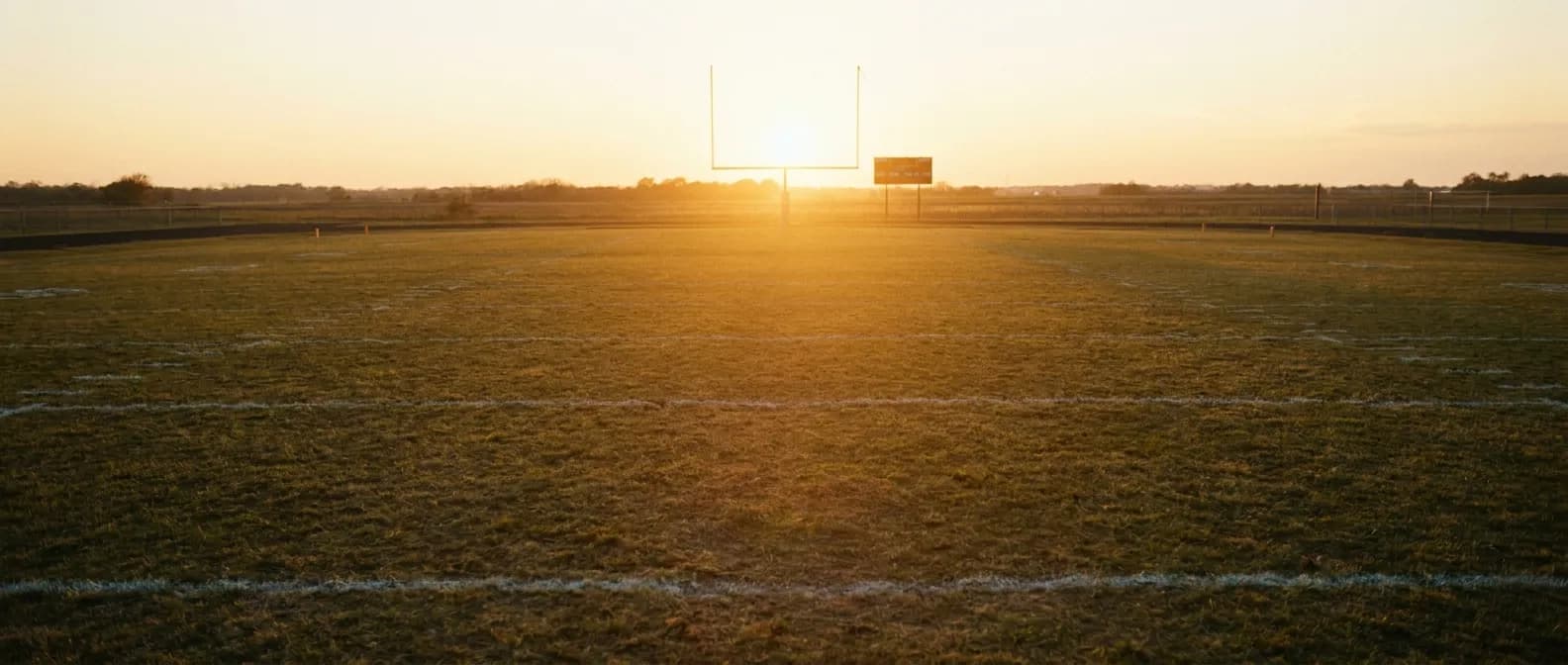 Sun setting over an empty football field with yard lines stretching into warm haze