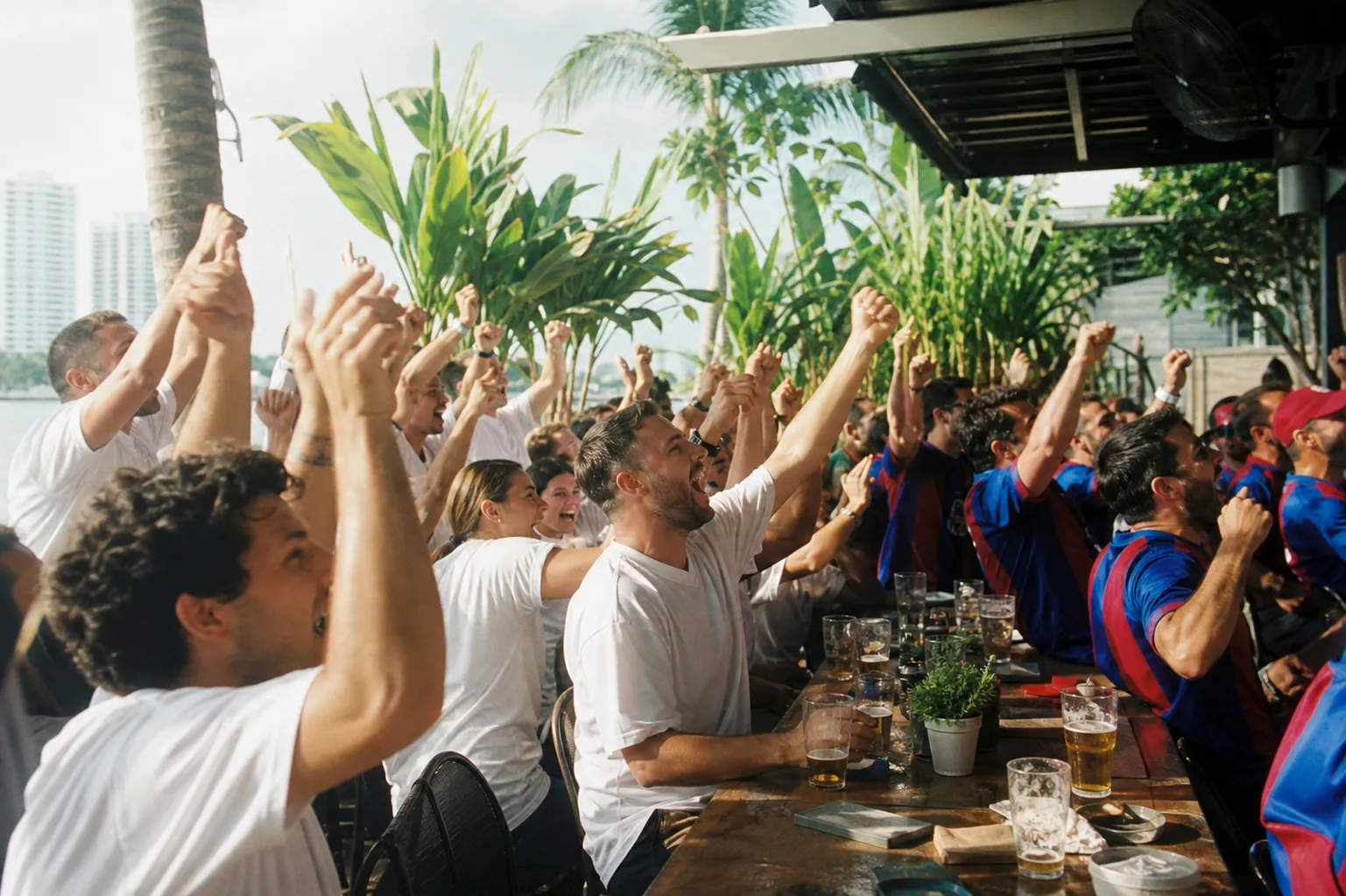 Two groups of soccer fans in plain white and in blue-and-red shirts sharing a long outdoor table in Miami, drinks raised toward an off-camera screen