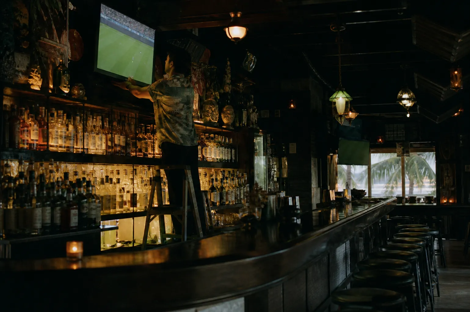 A bartender adjusting a television above rows of bottles in a dim Miami bar, the screen showing a soccer pitch from a distance