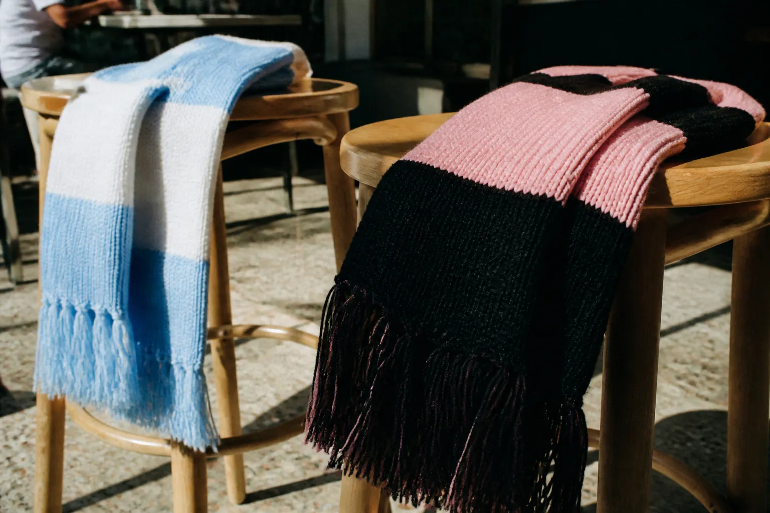 Close-up of two soccer scarves in contrasting colors draped over adjacent bar stools, sunlight cutting across a terrazzo floor