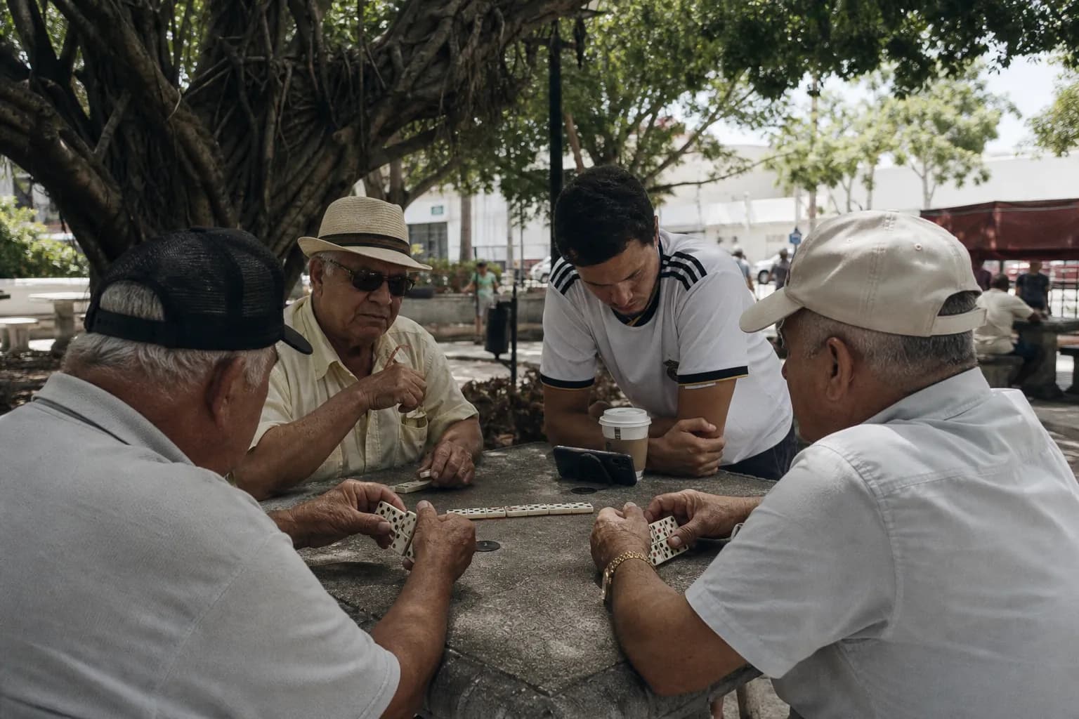 Older men playing dominoes at a shaded park table in Little Havana while a younger man in a soccer jersey watches a phone propped against a coffee cup