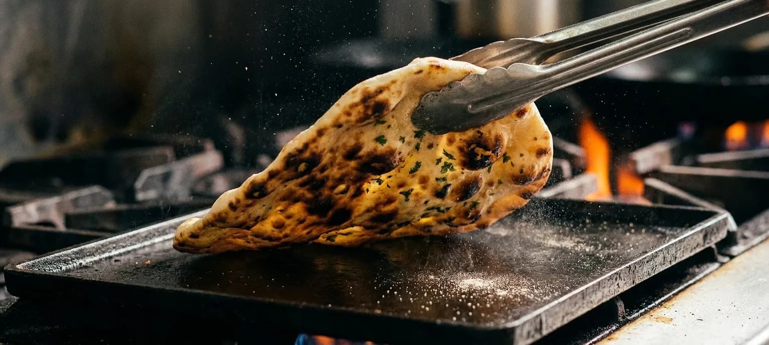 A blistered charred flatbread being lifted from a hot metal surface with tongs, flour dust in the air