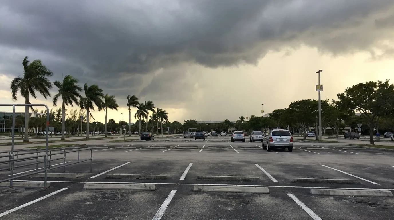 Afternoon storm clouds gathering over a Miami parking lot with scattered vehicles and palm trees swaying