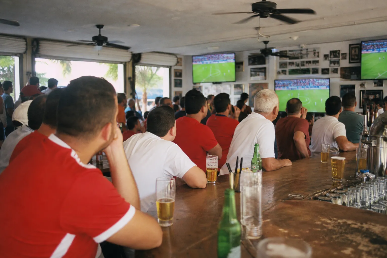 A crowded Miami sports bar in daylight with ceiling fans, several screens showing soccer at a distance, and fans in unmarked red and white shirts leaning over the counter
