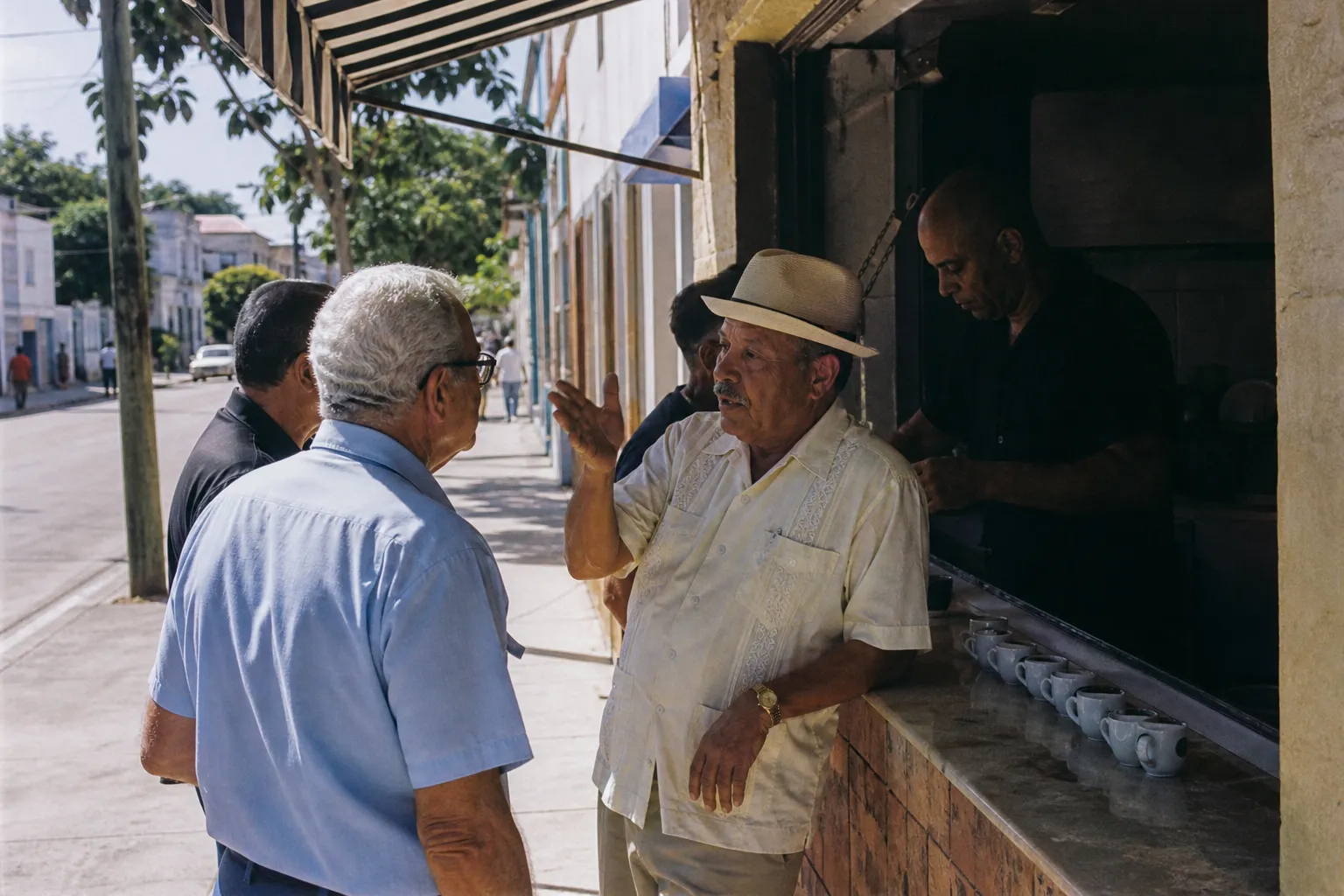 A walk-up cafecito window on a Little Havana sidewalk with small espresso cups on the counter and two men in guayaberas talking in the shade of an awning