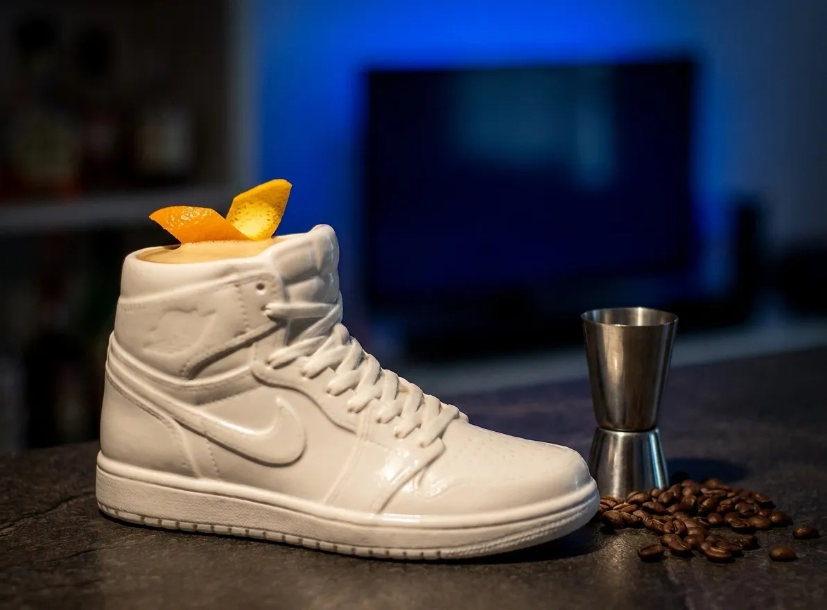 An unusual ceramic cocktail vessel shaped like a high-top trainer, filled with a dark drink and a single orange twist, on a bar counter