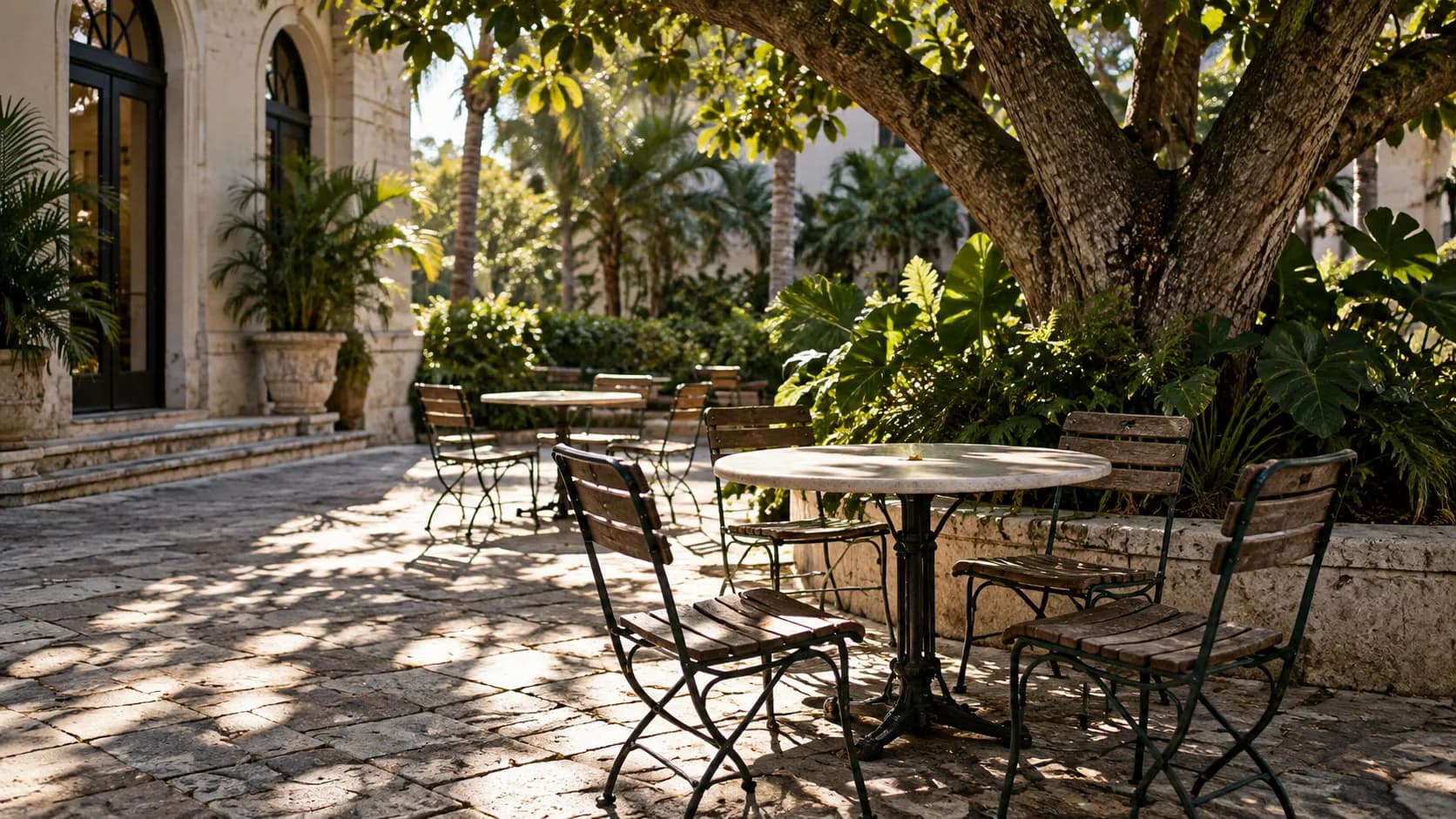 Empty metal-and-wood chairs beneath a broad leafy tree on patterned paving, with late sunlight casting tropical shadows across the quiet courtyard.