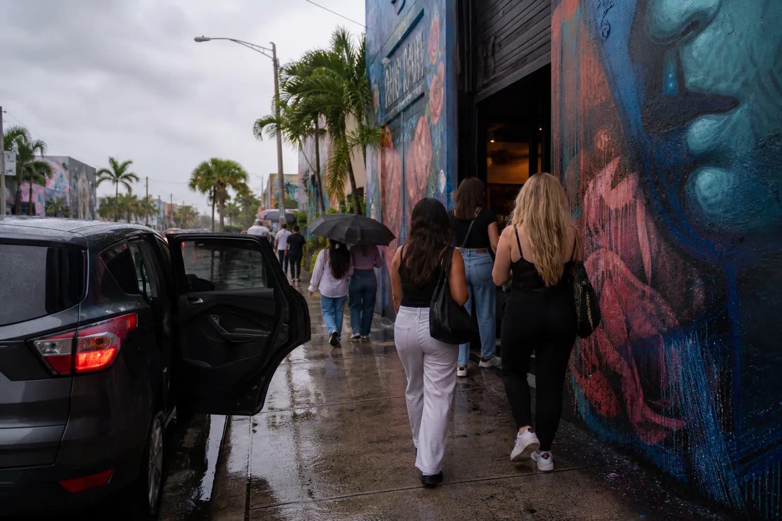 Mural-lined Wynwood street on a humid afternoon with a rideshare pull-up and people heading to an indoor class.