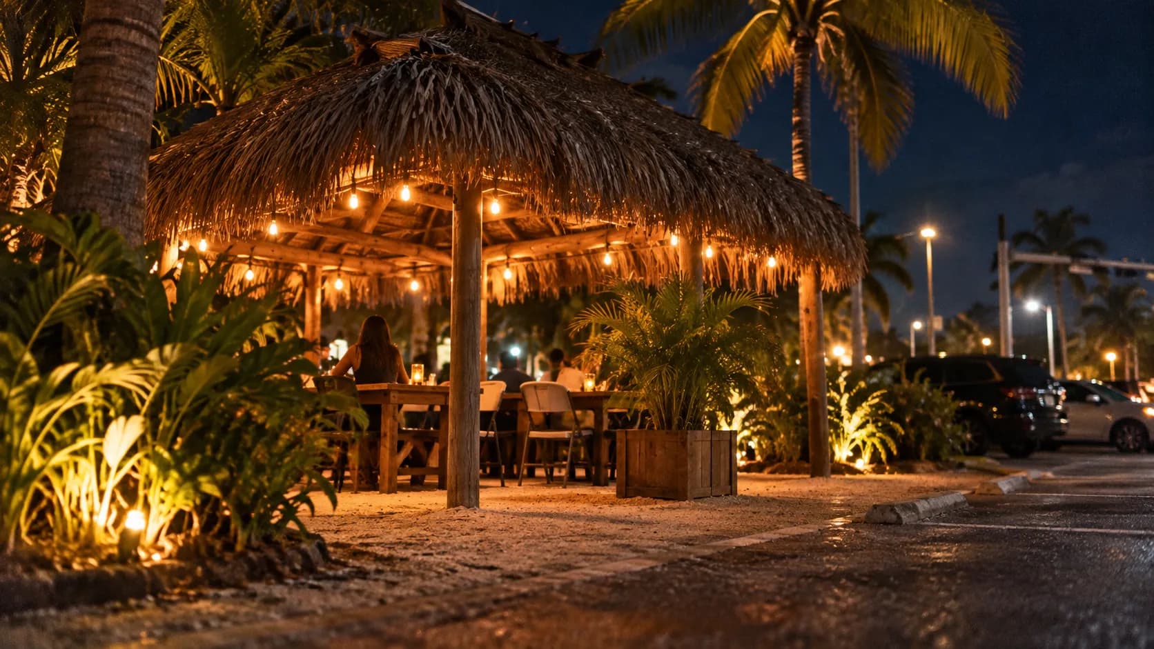 Night view of Chef Creole’s open-air palm-frond hut, warm bulbs, sandy ground, tropical greenery and parked cars beneath a dark Miami sky.