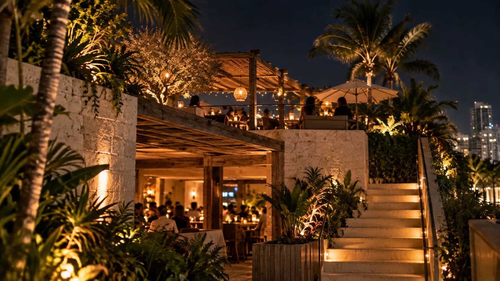 Street-level night view towards MILA’s rooftop terrace, with pale stone, reclaimed wood, dense greenery, amber light and open-air seating.