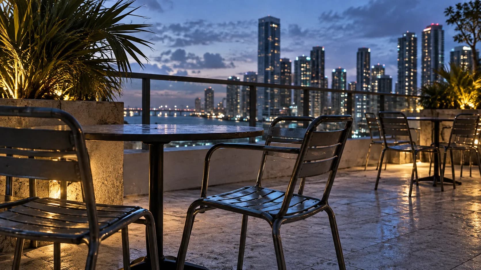 Close low-angle evening view of Julia & Henry's rooftop terrace, empty café chairs, pale stone, blue city light, and softened downtown skyline.