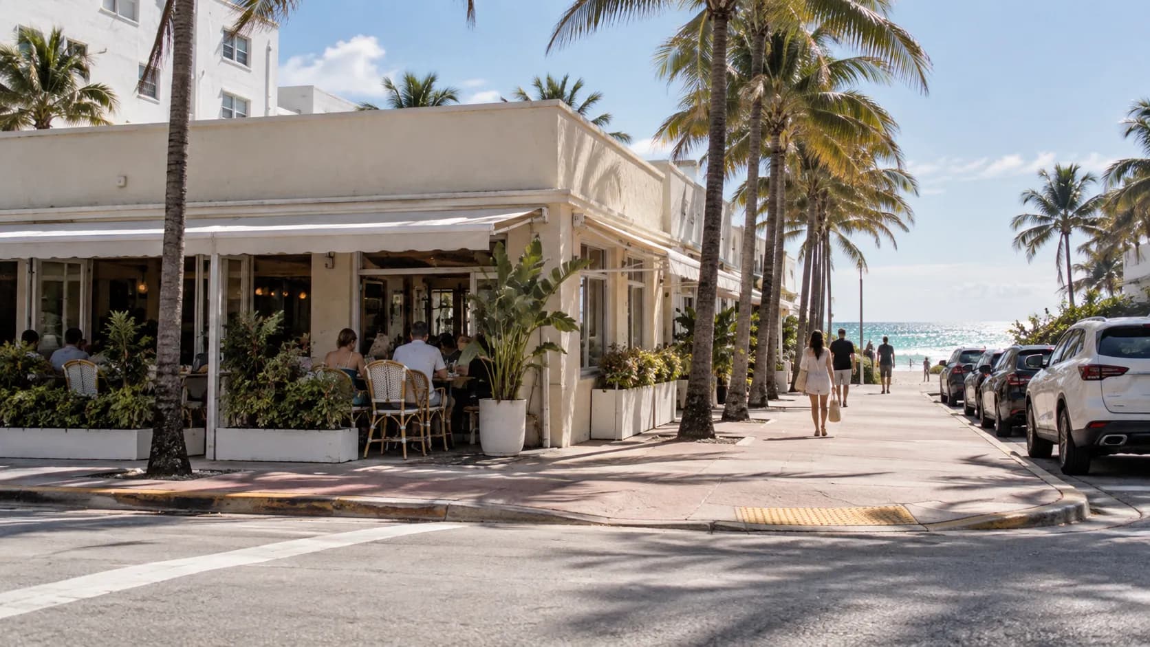 A wide daytime view of Manolo’s low corner restaurant on Collins Avenue, with pale stucco, palms, parked cars and bright North Beach light.
