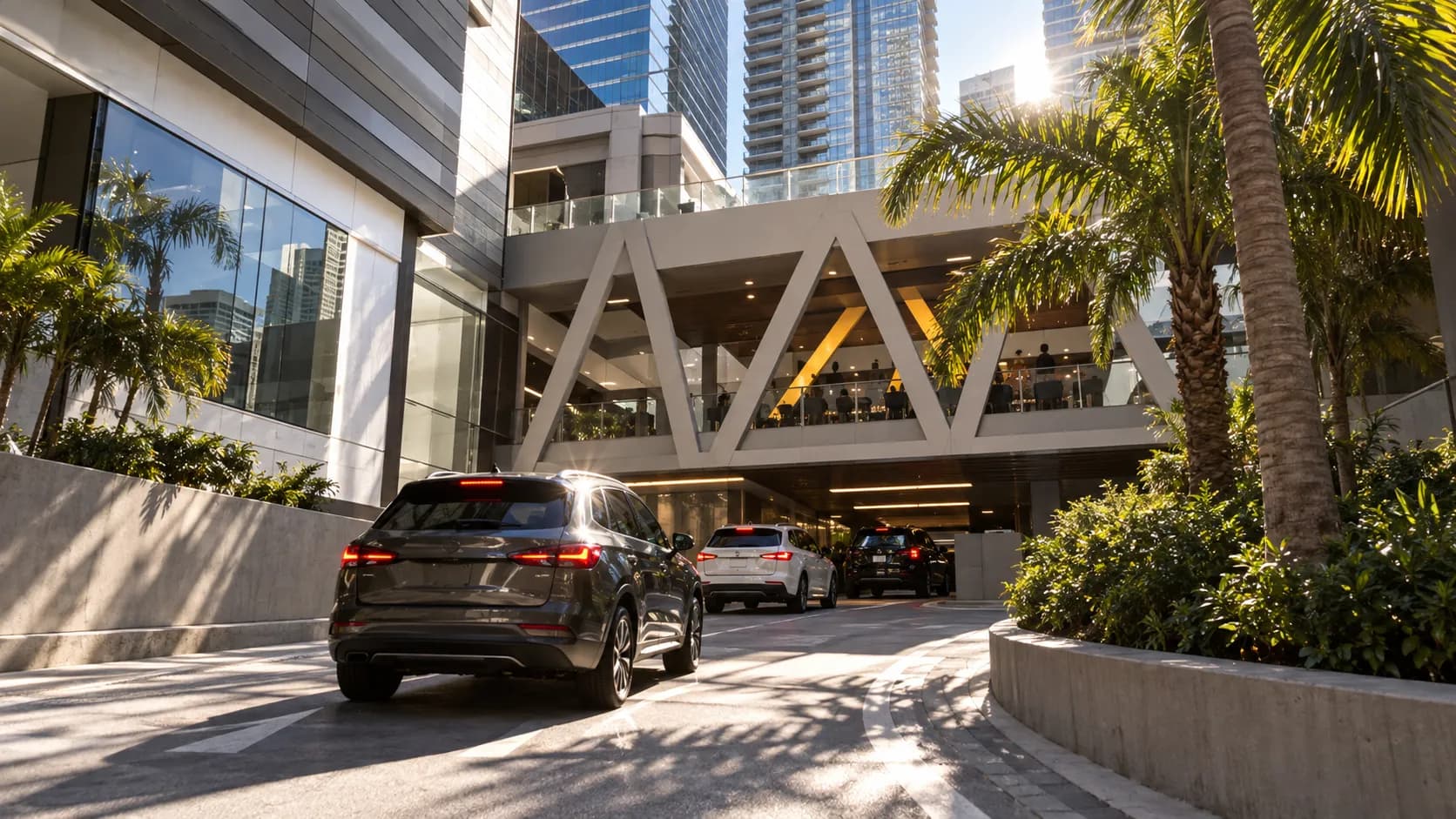 Cars turn into the Brickell World Plaza driveway at late afternoon, with hard sun on concrete and glass towers beyond the garage entrance.