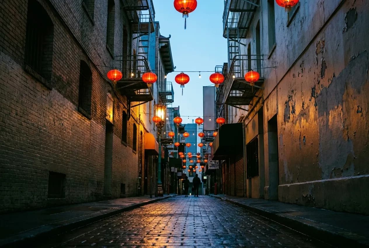 A narrow Chinatown alley at dawn with red lanterns overhead and wet pavement reflecting morning light.