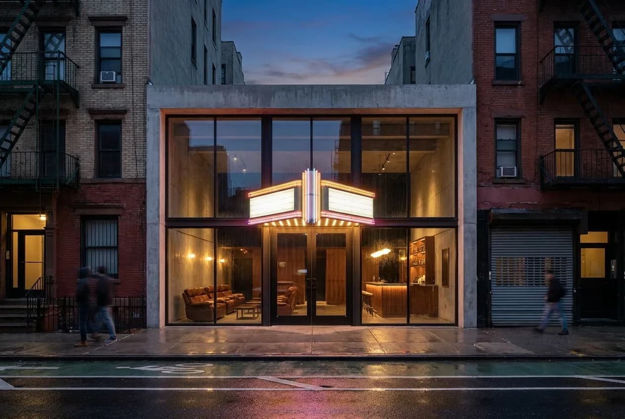 Narrow Ludlow Street entrance to a minimalist cinema with vintage marquee lighting and glass doors opening onto a concrete lobby at dusk in Lower East Side