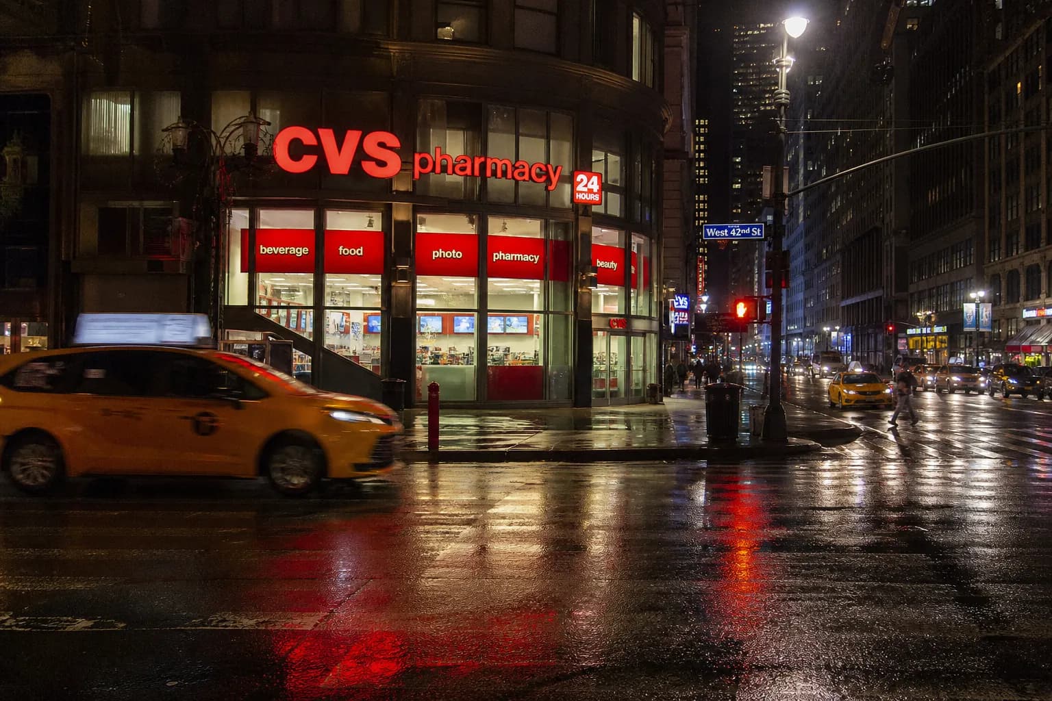 The bright red and white neon sign of a CVS storefront glowing on a wet Manhattan street corner at night.