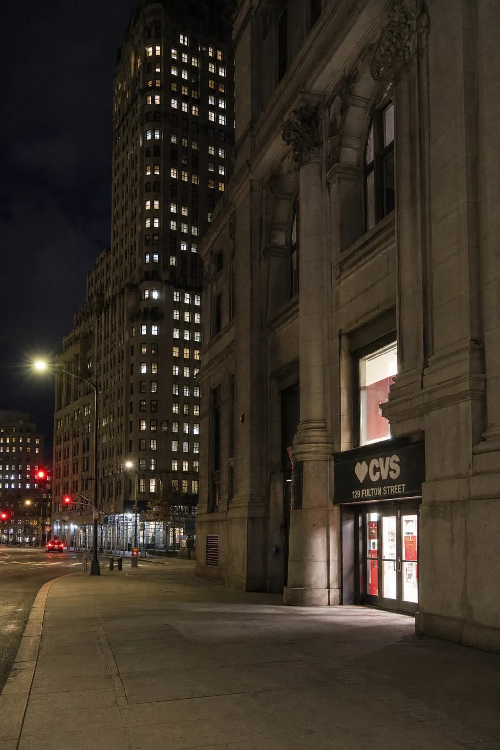 Looking up at a CVS entrance on Fulton Street in Lower Manhattan surrounded by tall brick buildings at night.