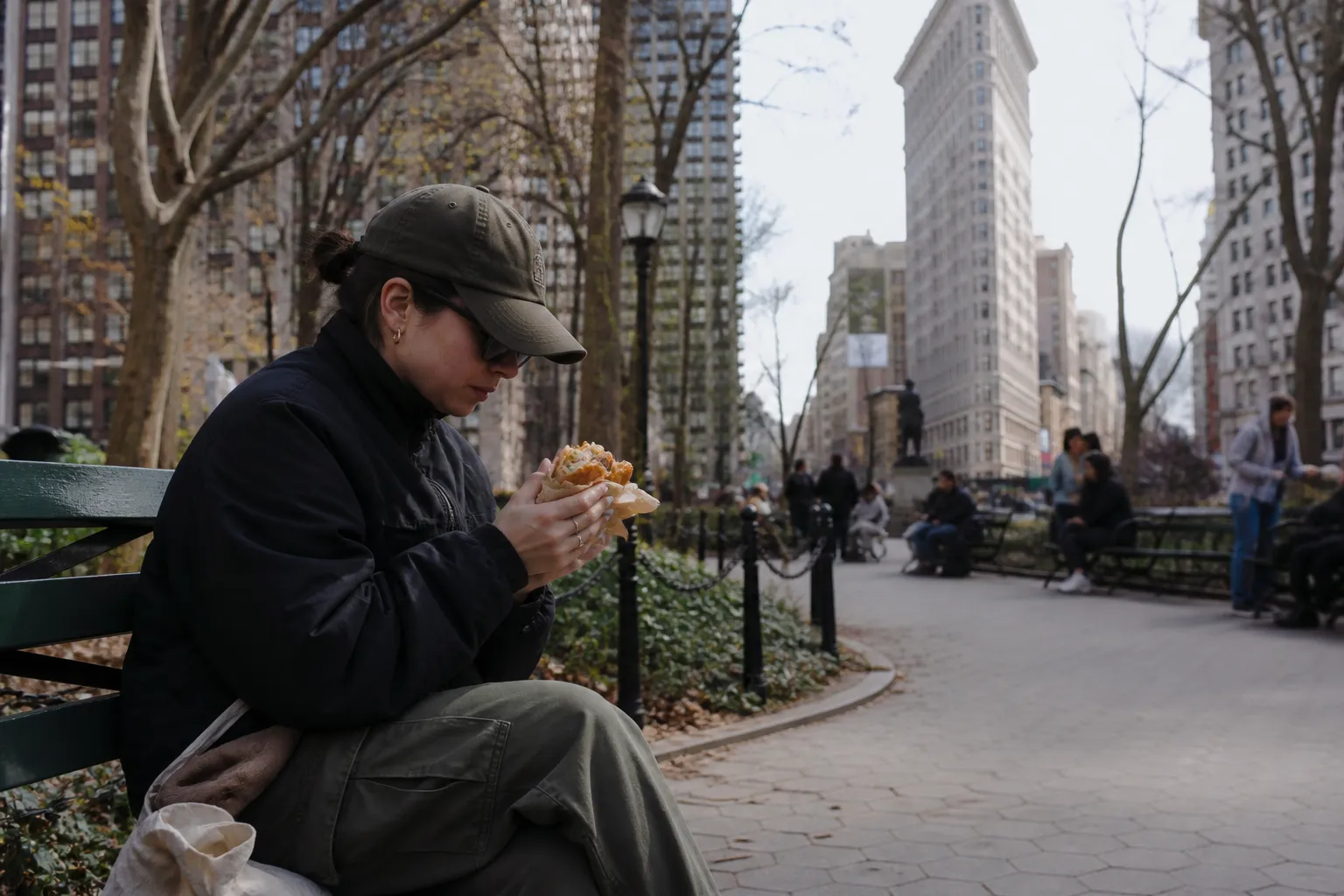 A person holding a wrapped chicken sandwich in hand, sitting on a park bench in Madison Square Park with NYC architecture in the background.