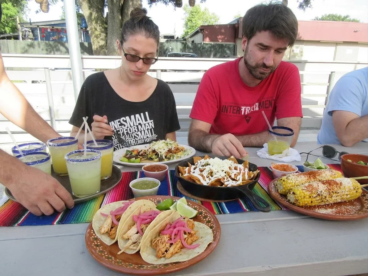 A brunch table covered in shared Mexican plates, tacos and chilaquiles, with a tray of margaritas arriving from the side