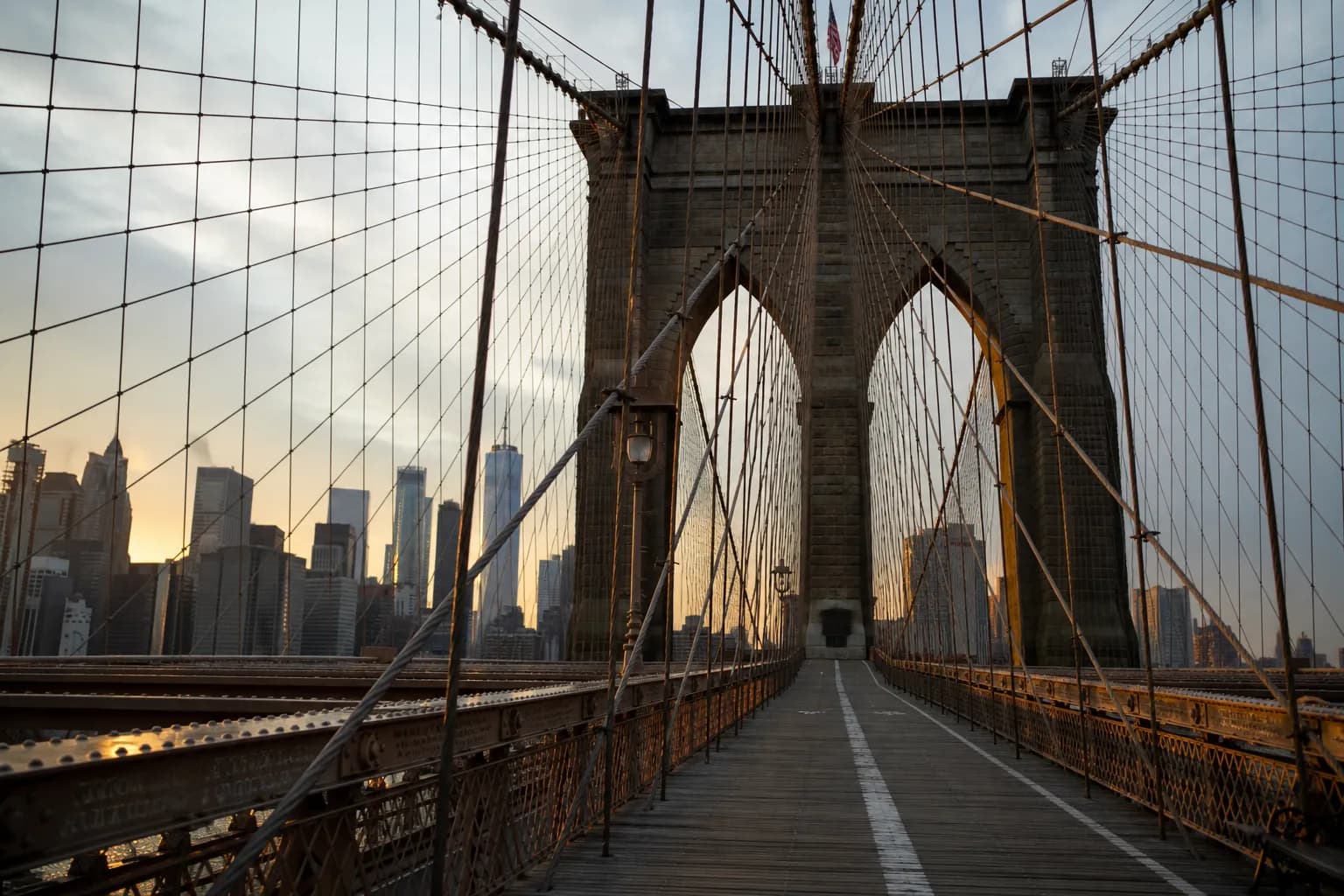 A wide-angle shot of the Brooklyn Bridge pedestrian path at sunrise looking toward Manhattan