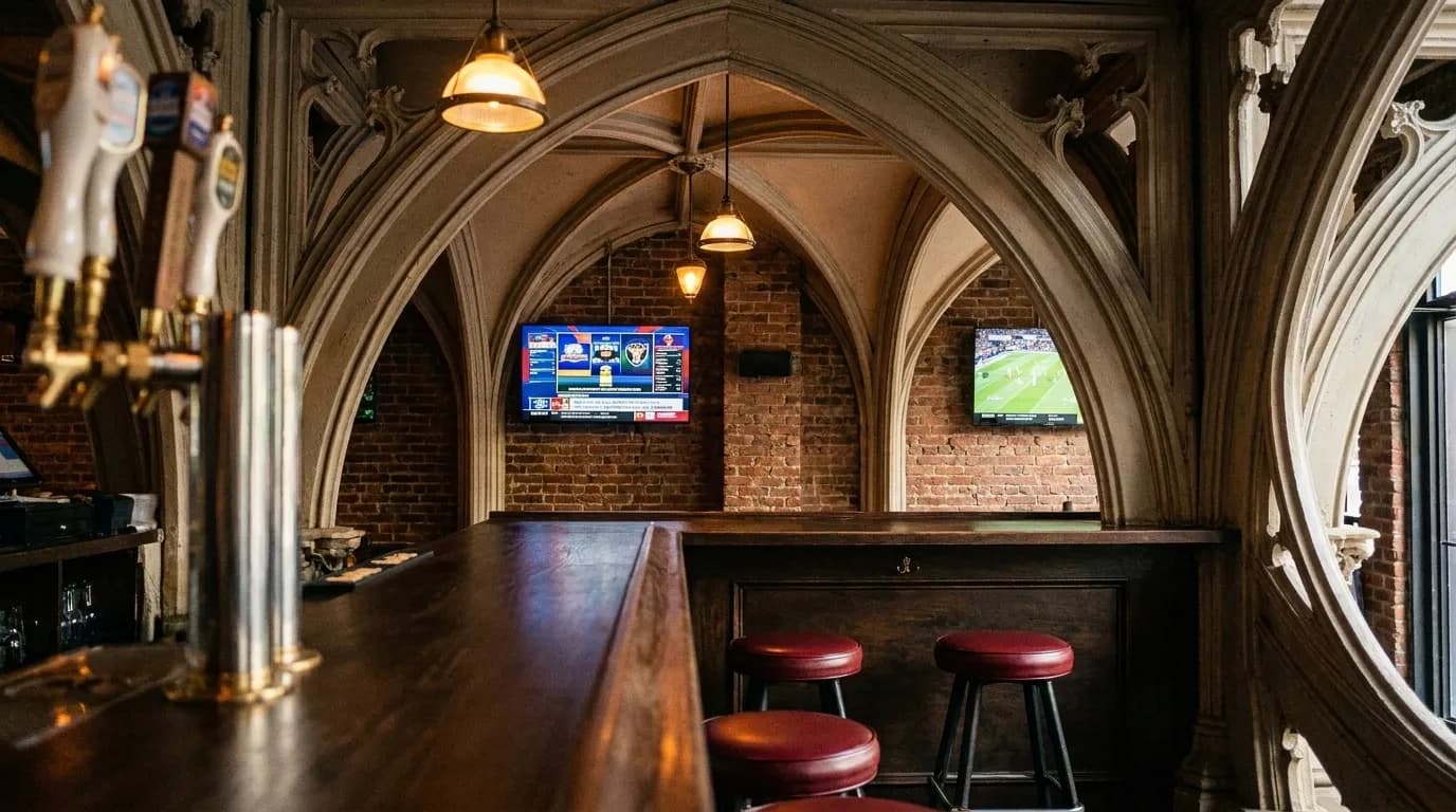 Interior of a small Brooklyn bar with flat-screen TVs on exposed brick, wooden bartop, and empty stools in warm lighting