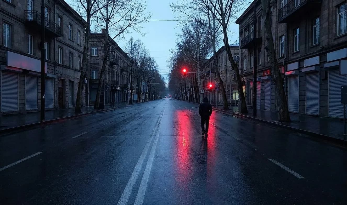 A city street at four in the morning with wet asphalt reflecting a traffic light and shuttered frontages under a lightening sky