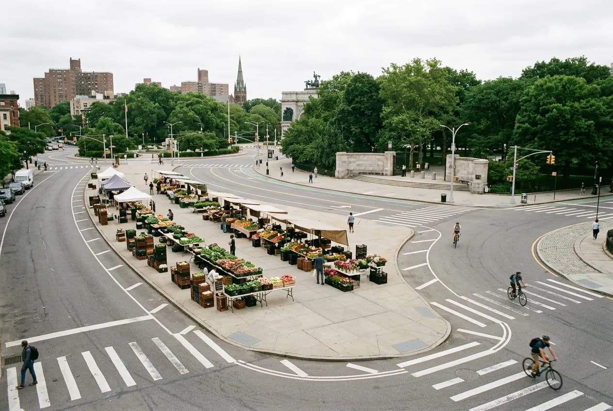 Outdoor farmers market stalls set up along a paved plaza at the edge of a park, morning light, city trees behind