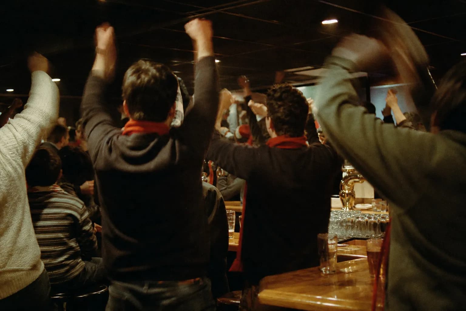 A crowd of soccer fans in plain scarves rising off their stools mid-cheer in a dim bar, arms up toward an unseen screen