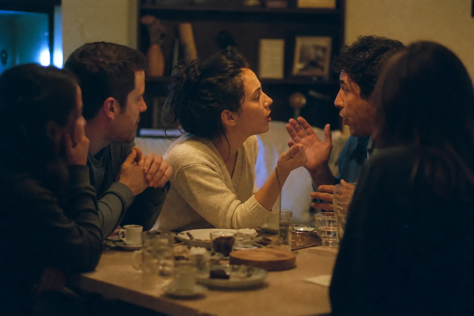 Friends leaning over a table of small plates in animated conversation, lit by the glow of an out-of-frame television