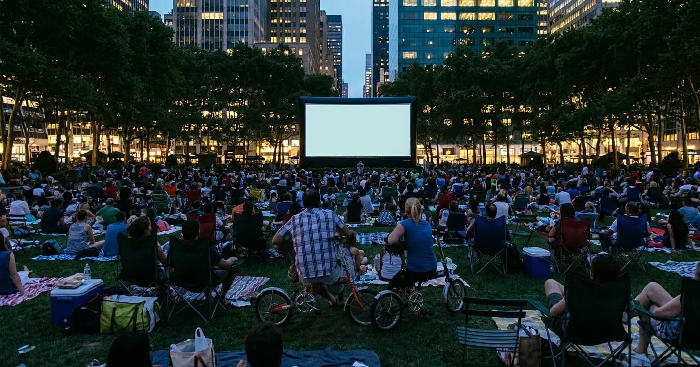 A midtown Manhattan park lawn at dusk during a free screening, silhouetted crowds on blankets facing a glowing blank screen with lit towers behind