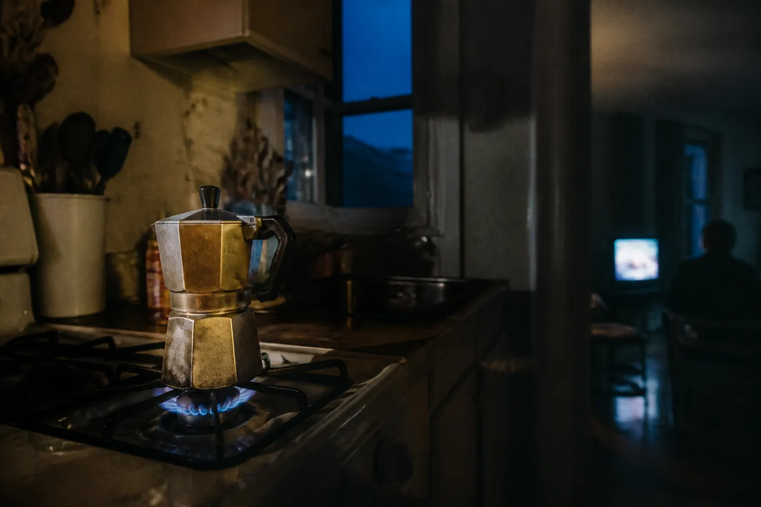 A stovetop moka pot steaming on a burner in a dim pre-dawn New York kitchen, a television glowing faintly in the next room