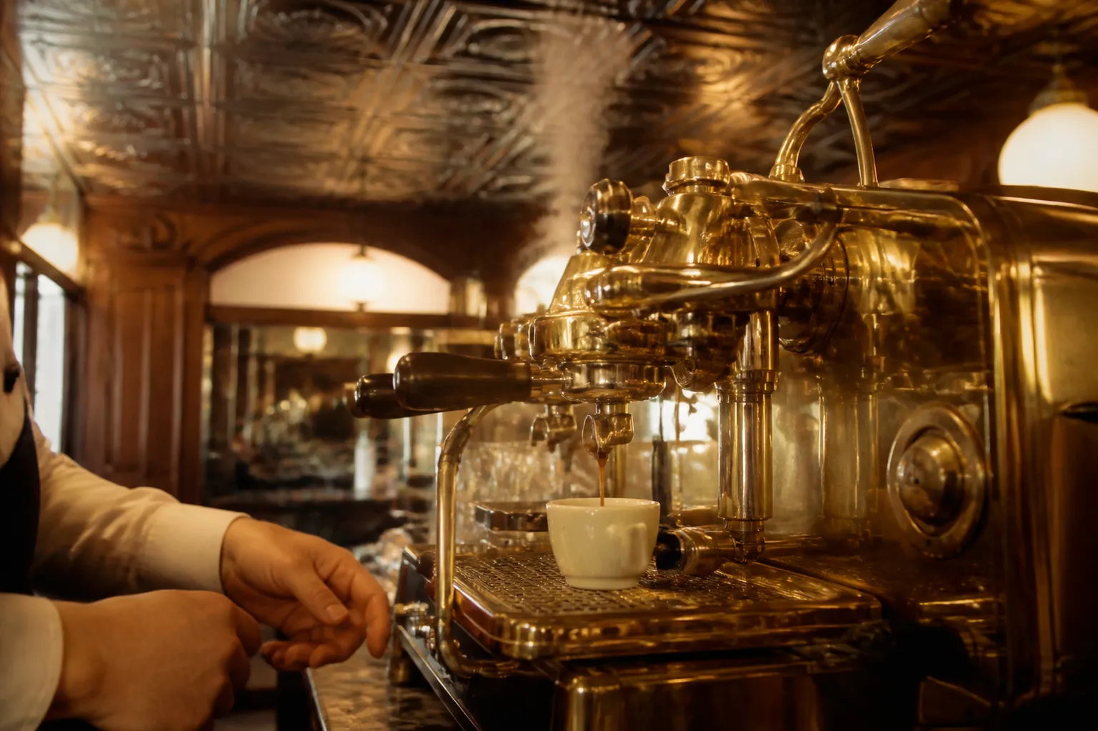 A brass espresso machine pulling a shot into a small white cup at an old-world caffe counter, steam curling in warm lamplight