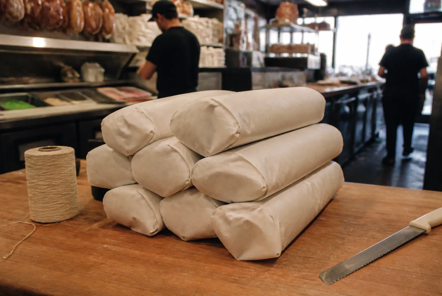 Foot-long hero sandwiches wrapped in white butcher paper stacked on a deli counter beside a spool of twine