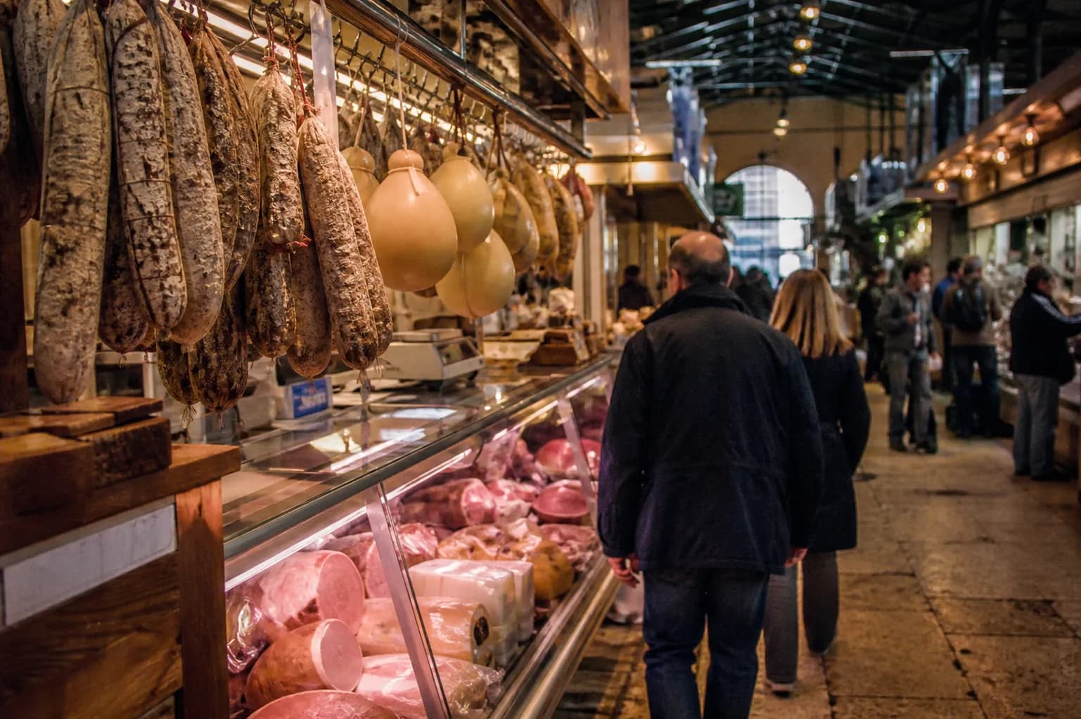 Inside a covered Italian market with sopressata and provolone hanging above a busy deli counter, shoppers blurred in motion
