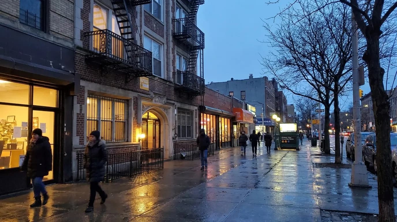 Franklin Avenue in Crown Heights at dusk, storefronts lit, subway entrance visible, pedestrians on sidewalk