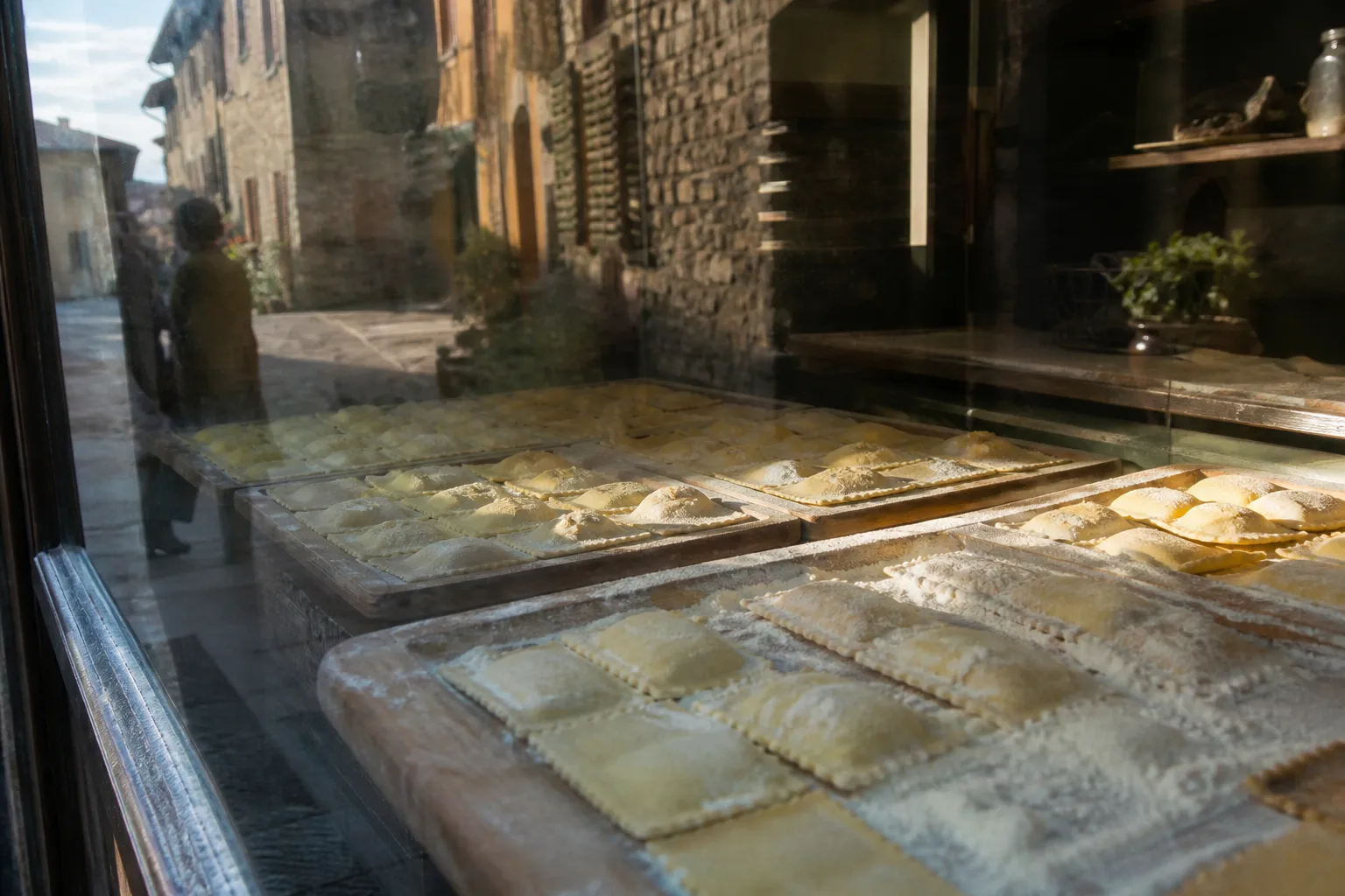 Fresh ravioli arranged in rows on floured wooden trays behind a pasta shop window, afternoon light raking across the flour dust