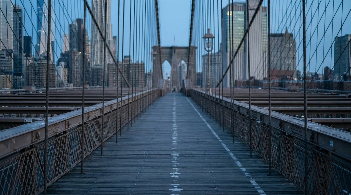 View along the Manhattan Bridge pedestrian walkway with steel cable railings and Brooklyn skyline in the distance during blue hour.
