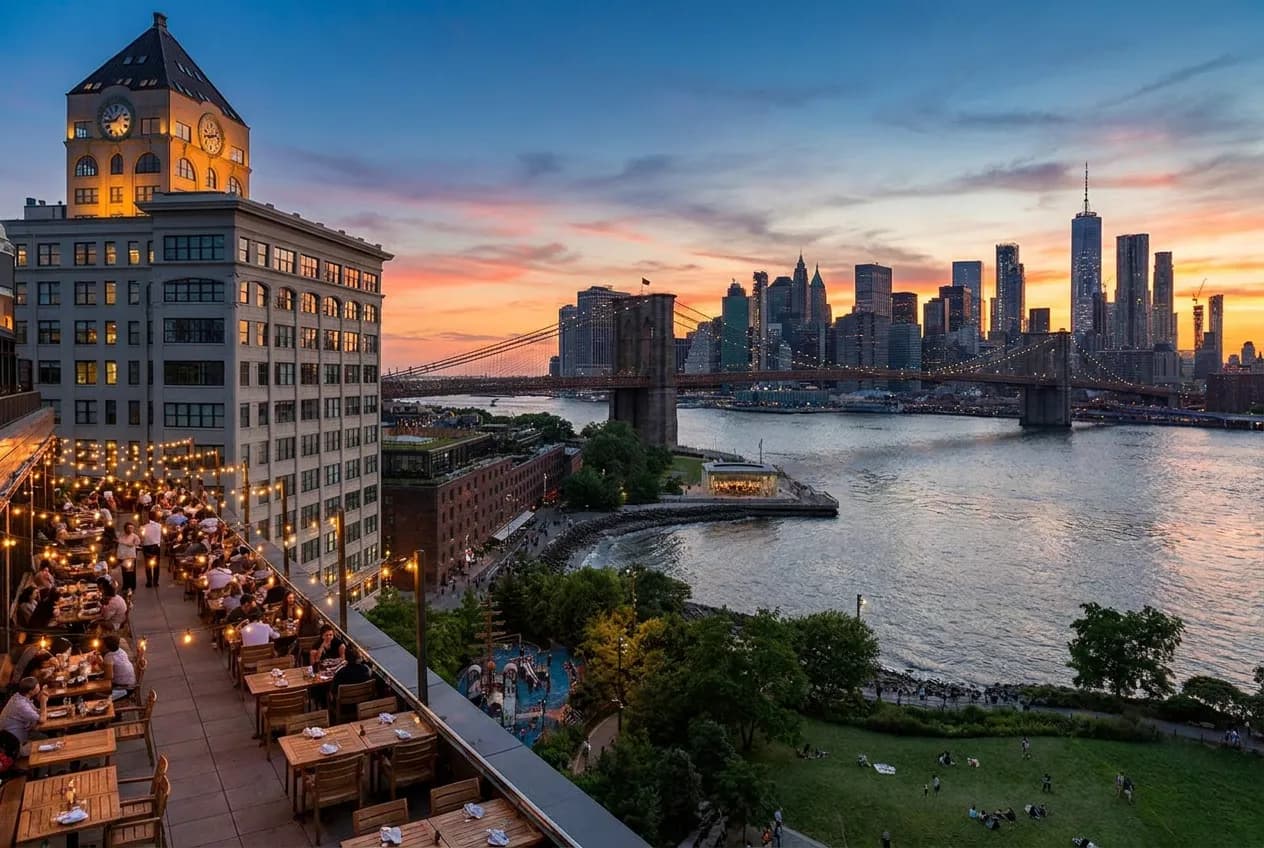 A waterfront restaurant terrace in Brooklyn Bridge Park at dusk with outdoor tables, string lights and the Manhattan skyline across the East River