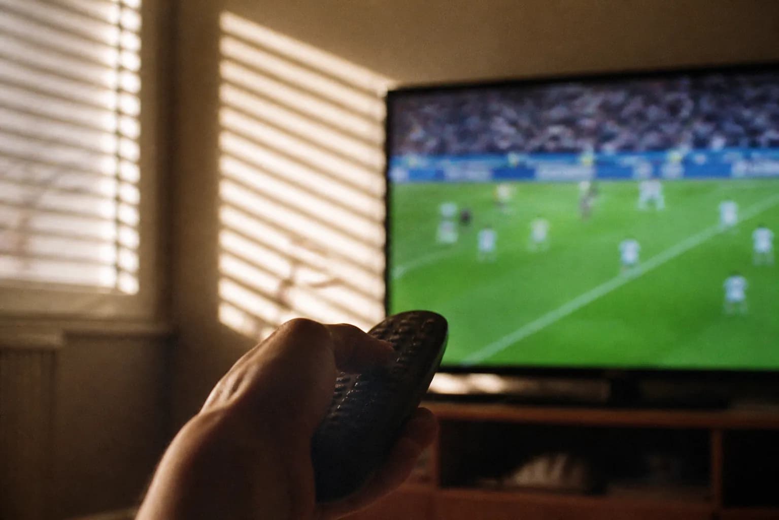 Close-up of a hand holding a television remote in front of an out-of-focus screen showing a green soccer pitch, morning light through blinds