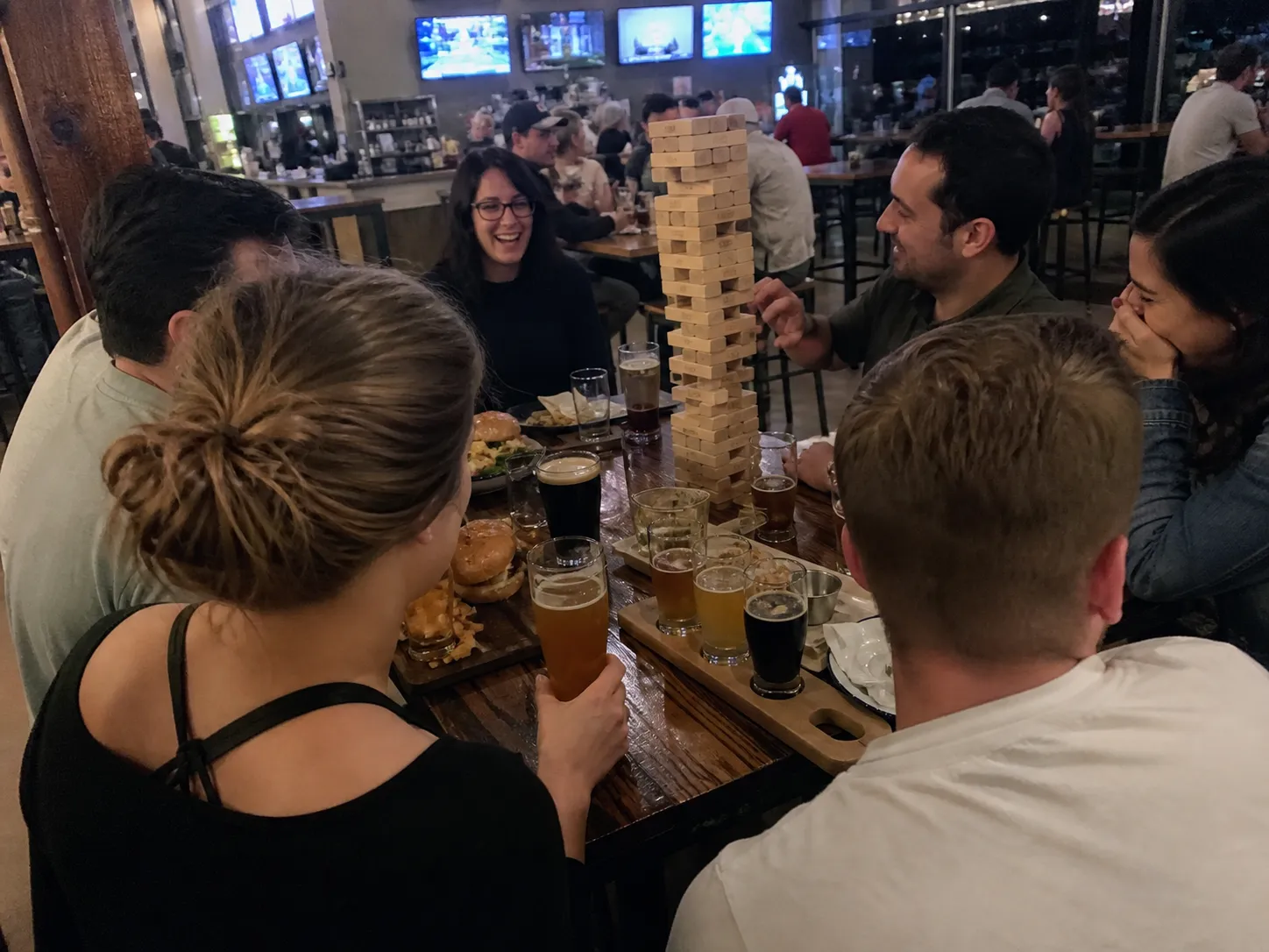 Friends around a communal beer-hall table in Manhattan with beer flights, burgers and a giant Jenga tower mid-game
