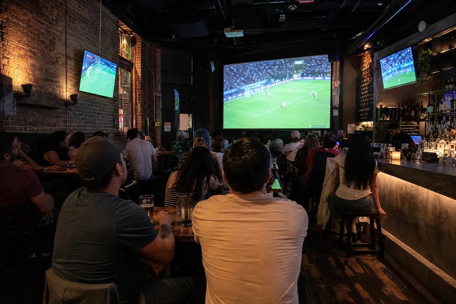 East Village sports bar with exposed brick, a large projection screen showing a soccer match and groups watching from wood tables
