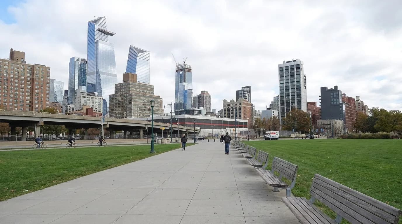 Wide view of East River Park promenade with open lawn, benches, and East River in the background under clear afternoon sky