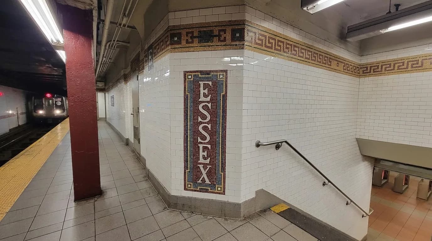 Interior of Essex Street subway station platform with tiled walls, metal columns, and train tracks under fluorescent light