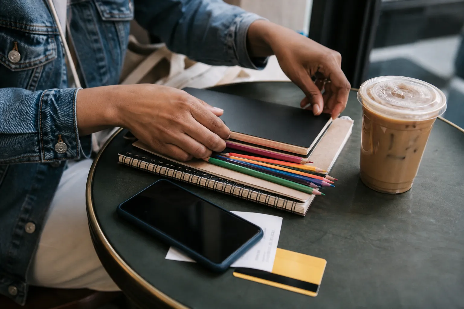 A cafe table with notebooks, colored pencils, iced coffee, a blank phone, and a plain transit-style card during a back-to-school errand break.
