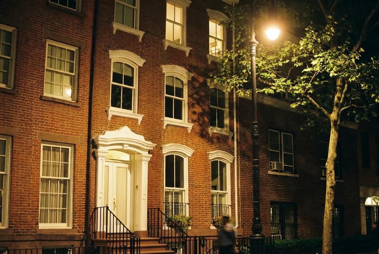 A quiet West Village street at night, lamp light through trees onto brick townhouses