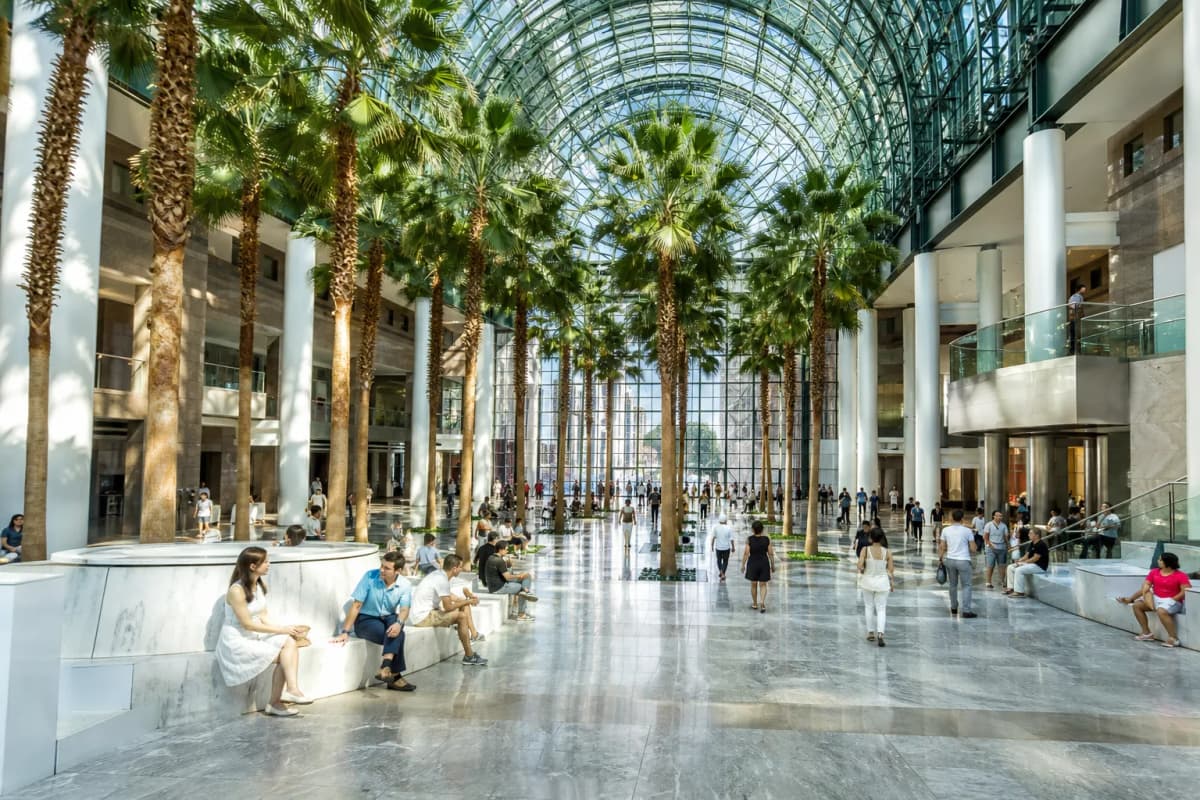 A bright Lower Manhattan indoor public concourse with glass roof, seating, and summer foot traffic