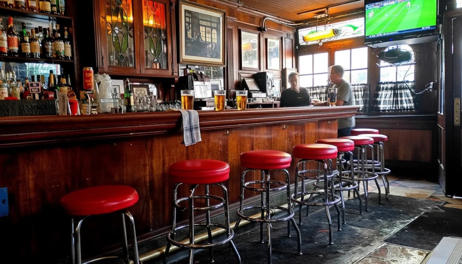 Classic Manhattan pub interior with a dark wood bar, red stools, pints of beer, and a game playing on a corner TV