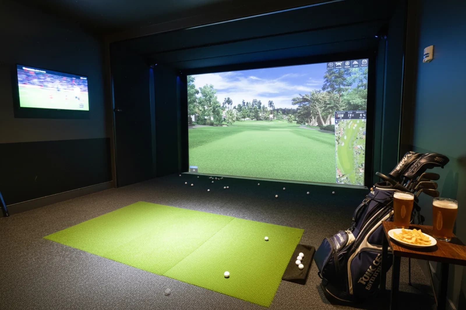 Golf simulator bay at T-Squared Social in Midtown, with a virtual fairway on the screen, a golf bag, and two beers waiting on a side table