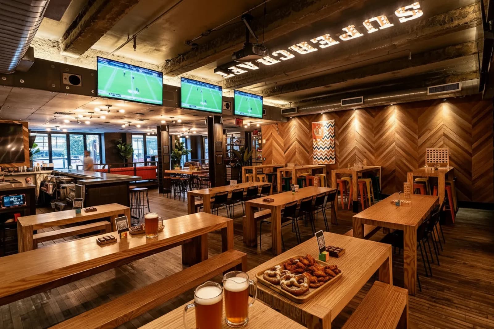 Communal wood tables at Clinton Hall 51 with beer mugs, a tray of pretzels and wings, and wall screens showing the game