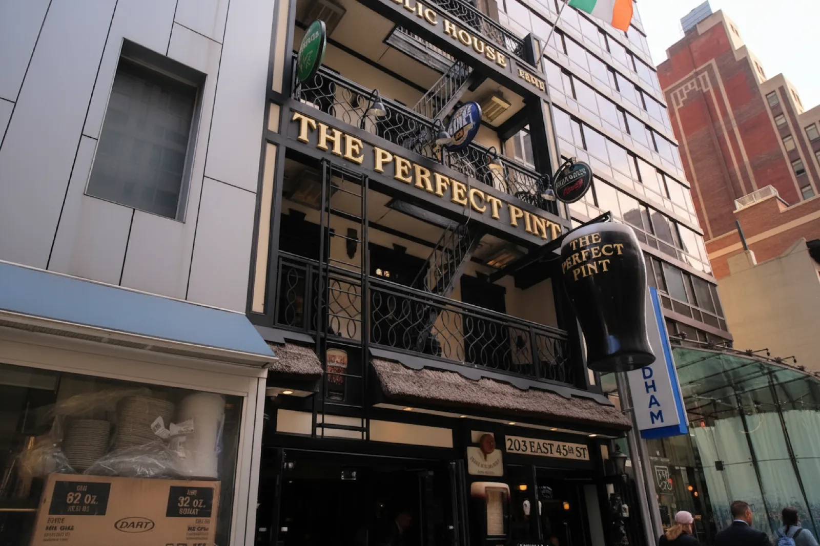 Street facade of The Perfect Pint East on East 45th Street, with its giant pint-glass sign, gold lettering band, balcony flags and thatched awning in early-evening light